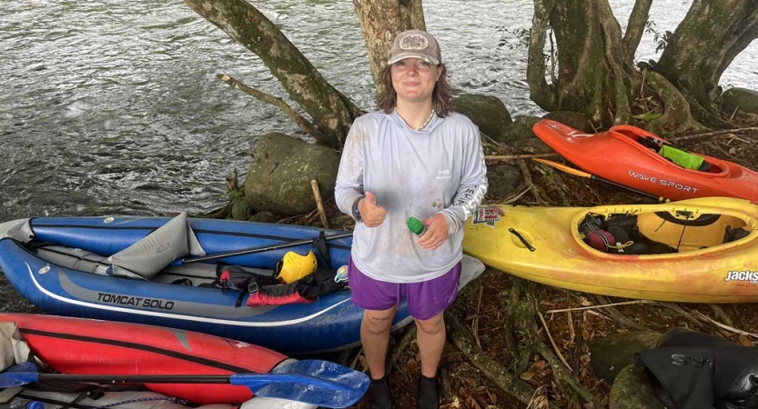 a person stands on the bank of a river, smiling and giving the camera a thumbs up. There are four watercraft on the bank nearby.